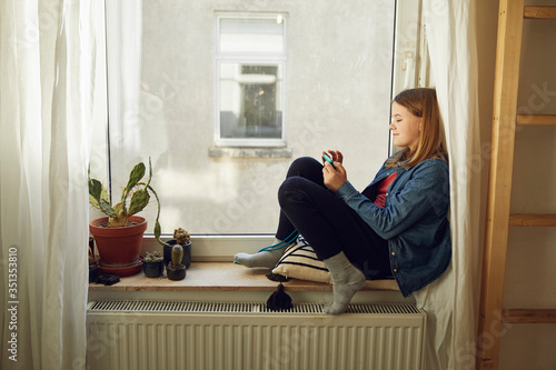 Girl sitting at the window at home using smartphone