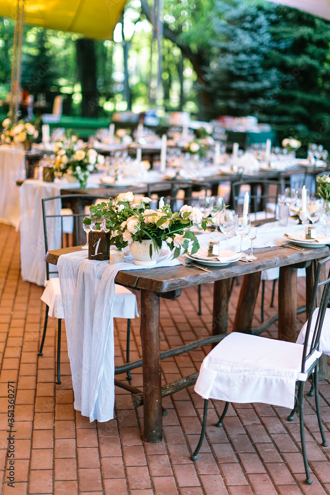 Obraz premium The rows of beautifully served wooden festive tables are arranged in a clear order, covered with fabric tablecloths, decorated with flower bouquets and filled with glasses and utensils.