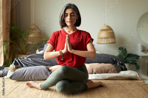 Fashionable young female with grayish hair sitting in gomukhasana or cow pose while practicing hatha yoga in bedroom after awakening, keeping eyes closed, pressing hands together in namaste photo