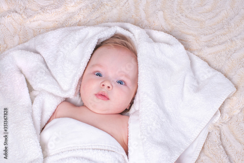 Newborn baby with light hair and blue eyes lies on bed on light blanket in white, soft, dry towel after bathing procedure. Concept of baby's daily routine and child hygiene