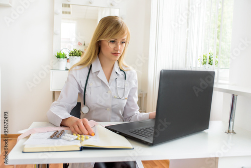 Female doctor working at office desk and smiling at camera, office interior.Online consultation, selection of treatment and examinations. Pills in the hands. Modern technology, telemedicine