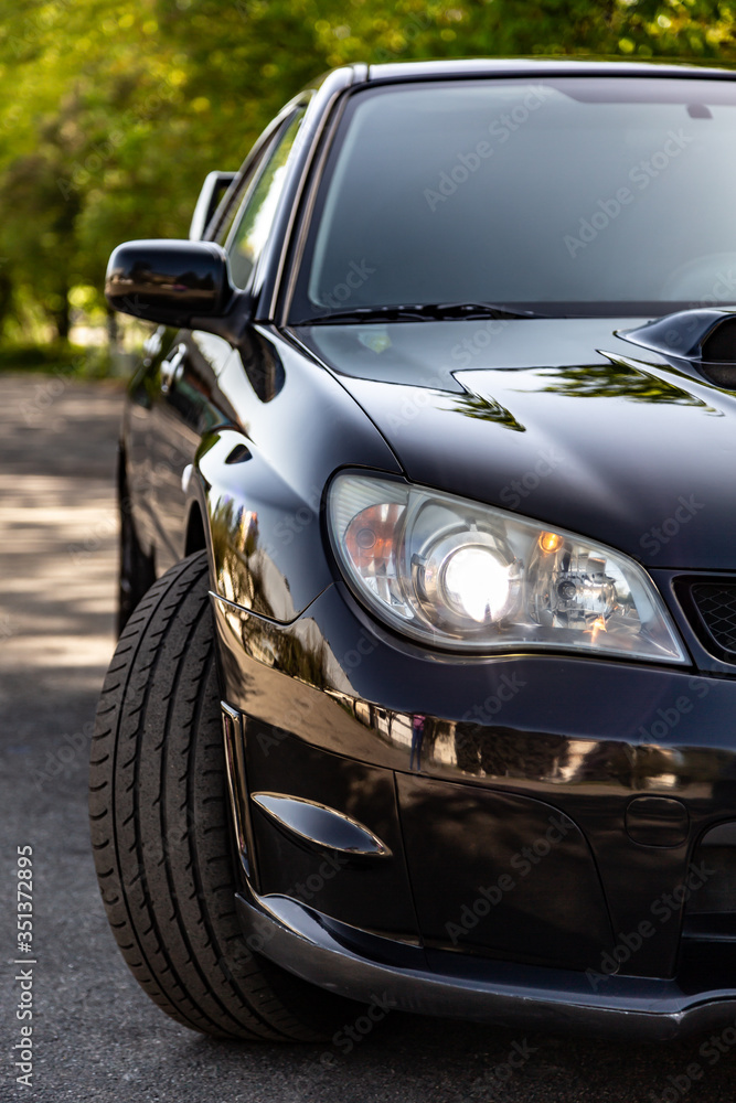 Black car in static on a sunny day. Front view half of the car - headlight and a turned wheel. Vertical orientation.