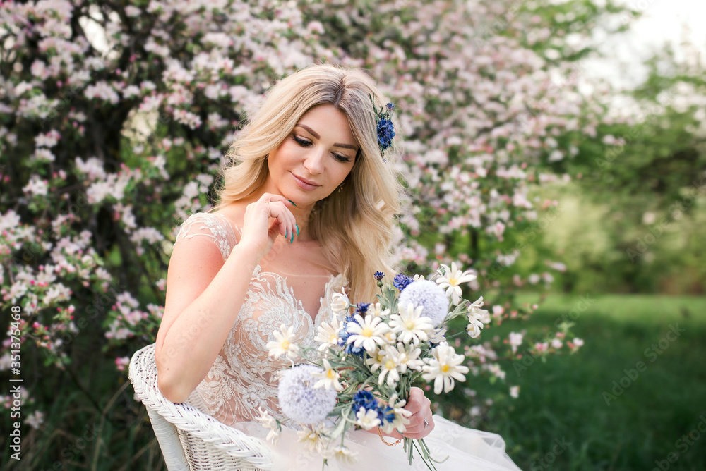 An attractive bride with a bouquet of wild flowers is sitting in a chair in a blooming garden An attractive bride with a bouquet of wild flowers is sitting in a chair in a blooming garden
