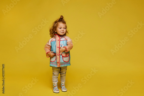 Stylish little girl in modish raincoat isolated at the yellow background. Child fashion. Copy space