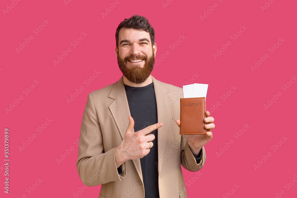 smiling bearded man is pointing at his passport and tickets he is holding over pink background. smiling bearded man is pointing at his passport and tickets he is holding over pink background.