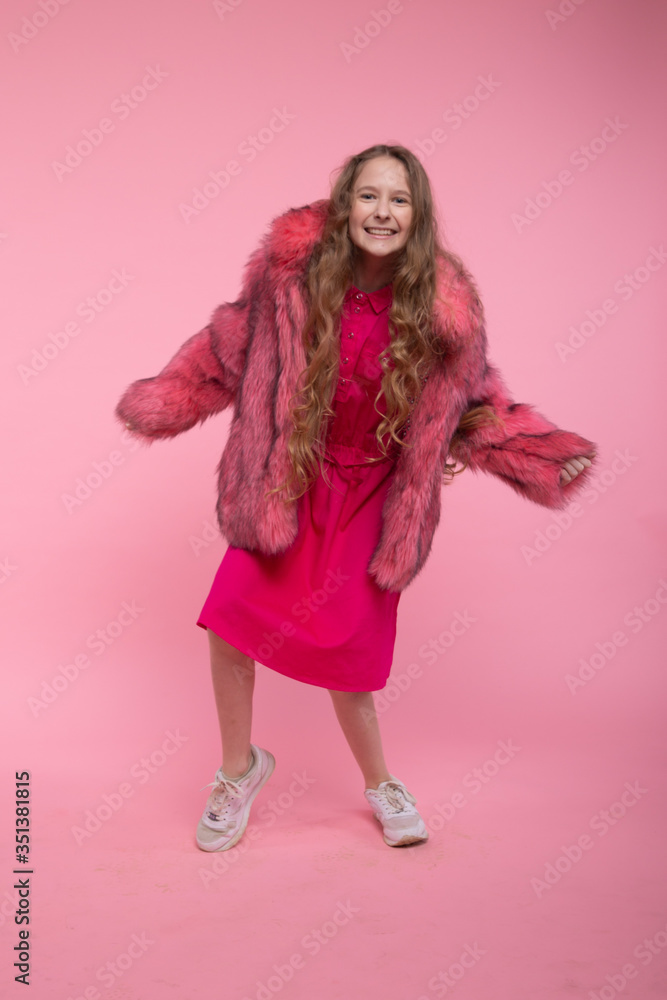 A cheerful girl in fashionable clothes, a light pink fur coat, a pink bright dress and white sneakers is dancing on a pink background. A cheerful girl in fashionable clothes, a light pink fur coat, a pink bright dress and white sneakers is dancing on a pink background.