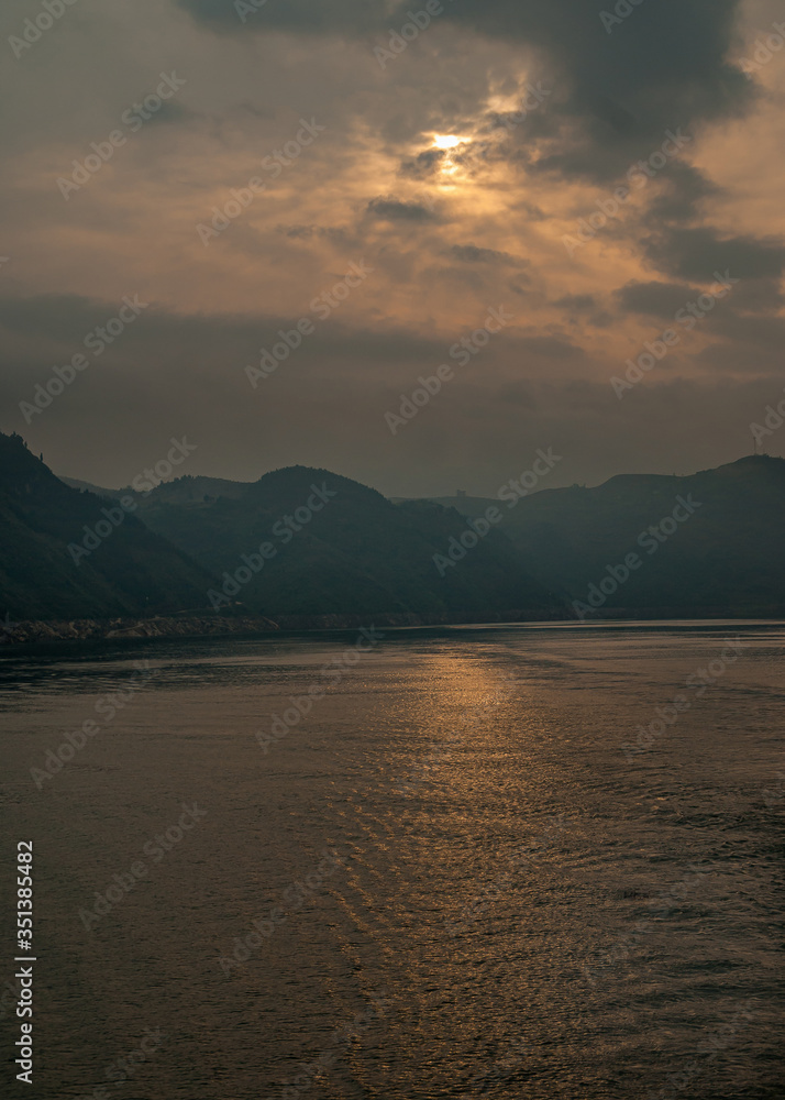 Guandukou, Hubei, China - May 7, 2010: Wu Gorge in Yangtze River. Early morning sun tries to break through dense cloudscape living golden line on dark water. Mountain range on horizon.