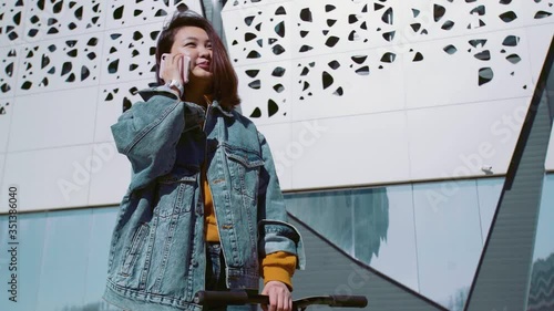 Young asian woman in a jeans jacket walks with the scooter and talks on the phone in a summer park photo