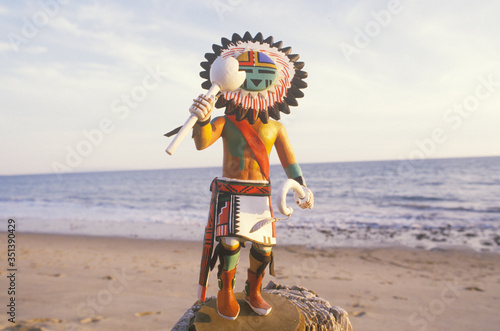фотографія Hopi Kachina doll holding objects on beach with ocean in the background