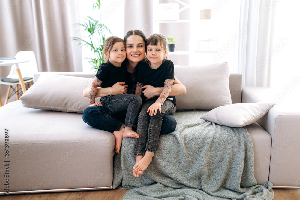 Mom and daughters. A wonderful portrait of a charming happy young mother and her two cute daughters. They are sitting on the sofa at home in casual stylish clothes hugging and smiling
