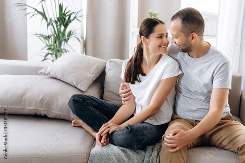 Happy couple in love in casual stylish wear sitting and hugging on a couch at their cozy apartment. Good moment. Relationship of spouses © Kateryna