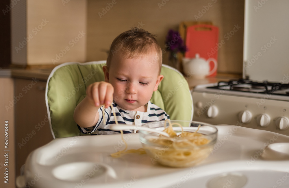 Funny boy eats spaghetti with his hands. A small boy is sitting on a high chair in the kitchen.