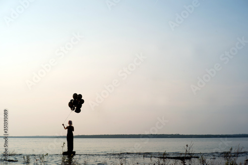 Woman on edge of frame in silhouette stands by the edge of a lake at sunset with black balloons about to let them go letting go of her past