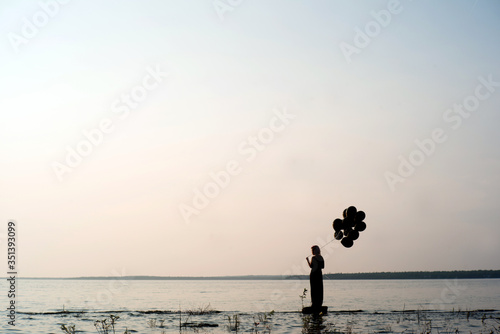Woman in silhouette stands by the edge of a lake at sunset with black balloons about to let them go letting go of her past