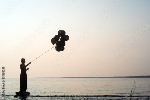 Silhouette of woman holding black balloons at sunset ready to let them go and fly away with lake and water behind her