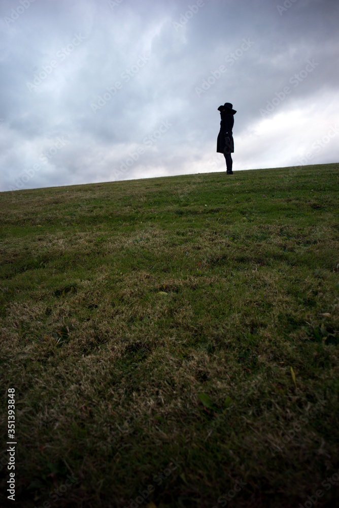 Lone woman wearing black hat and black jacket stands in the distance on top of hill alone and deep in thought against a cloudy sky