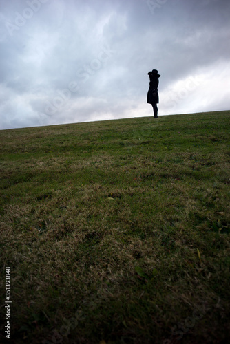 Lone woman wearing black hat and black jacket stands in the distance on top of hill alone and deep in thought against a cloudy sky
