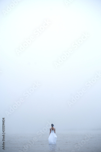 Woman in white dress and blue hair looks as if she is drifting by water surrounded by mist and fog 