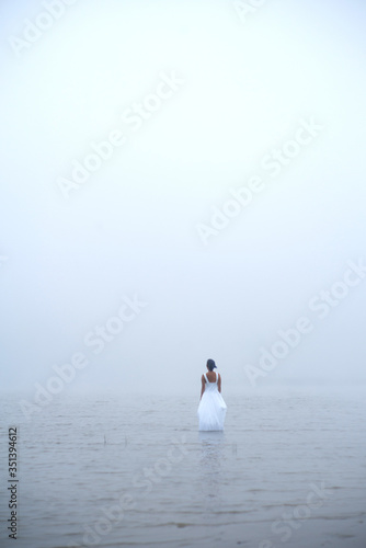 Woman in white dress and blue hair looks as if she is drifting by water surrounded by mist and fog 