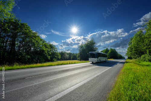 Obraz na płótnie White bus traveling on the asphalt road between the clearing and the forest under radiant sun w salonie