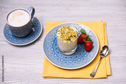 Pistachio parfait in a transparent glass cup on a blue plate with two strawberries, on a yellow napkin with a teaspoon. A cup of cappuccino stands on the side.