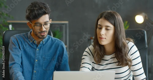 Muliethnic young guy and girl sitting at table together at laptop computer in office and working over new startup project. Mixed-races male and female company workers brainstorming and talking. photo