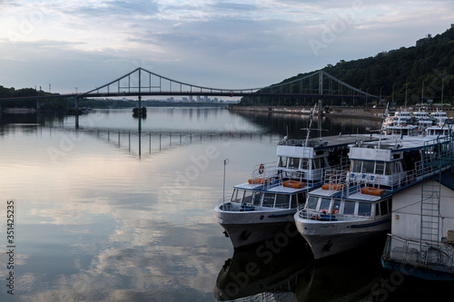 Boats near the pedestrian bridge on Dnipro river, Kyiv, Ukraine. Morning reflection in the water