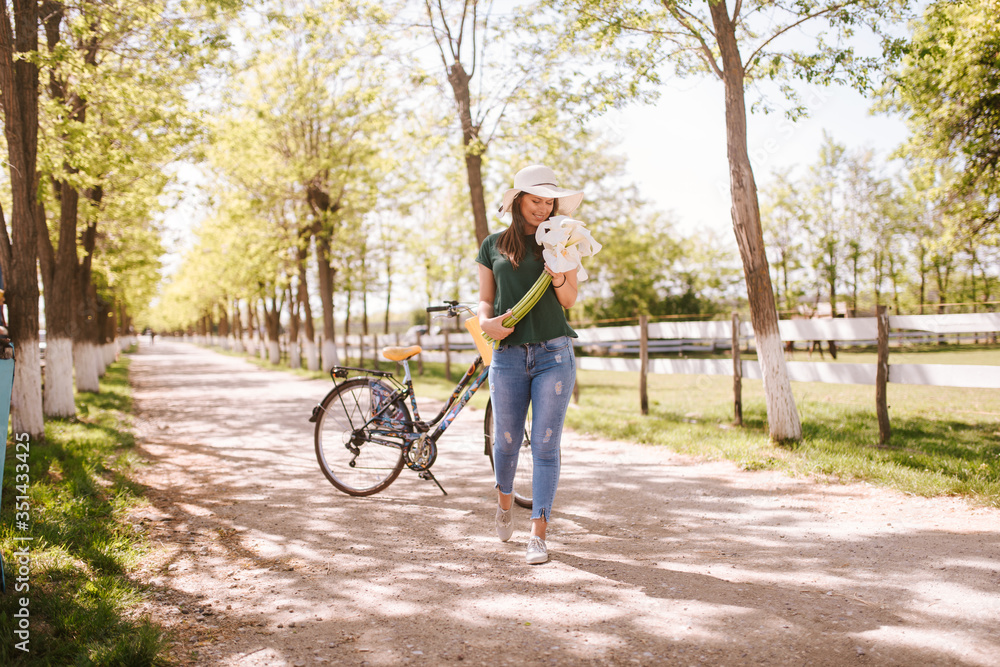 A beautiful young woman with a straw hat, bicycle and flowers is standing in the park. Life in a village