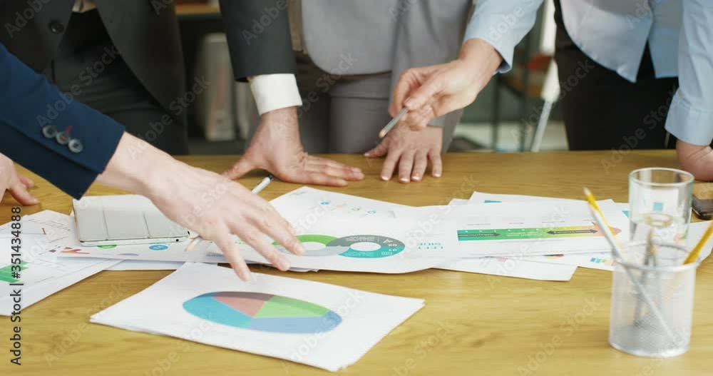 Close up of papers and documents with charts, graphics and diagrams lying on wooden desk in office. Business meeting and discussion concept. Mixed-races female and male hands. Multiethnic colleagues. Close up of papers and documents with charts, graphics and diagrams lying on wooden desk in office. Business meeting and discussion concept. Mixed-races female and male hands. Multiethnic colleagues.
