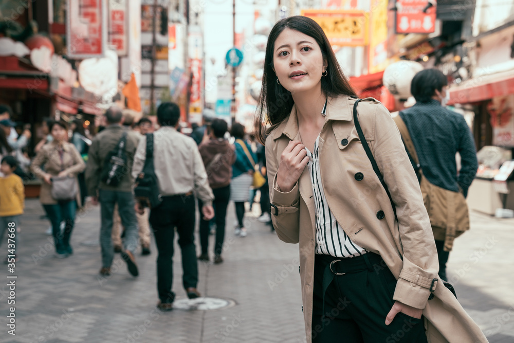Young attractive asian woman curiously looking around on street. smiling female standing with one hand in pocket sightseeing city osaka. happy girl in coat travel japan modern urban in spring season.