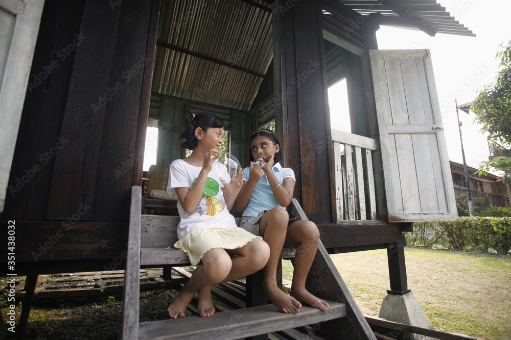 Girls sitting on stairs of wooden house blowing bubbles Girls sitting on stairs of wooden house blowing bubbles