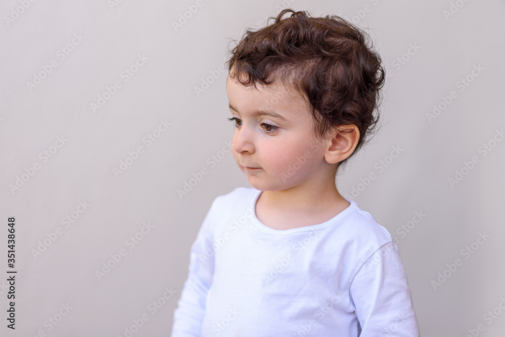 Naklejka premium Funny boy with curly hair in a white t-shirt on a grey background. Photo of adorable young happy kid looking away. Child of two years old smiles and laughs. Children's emotions.