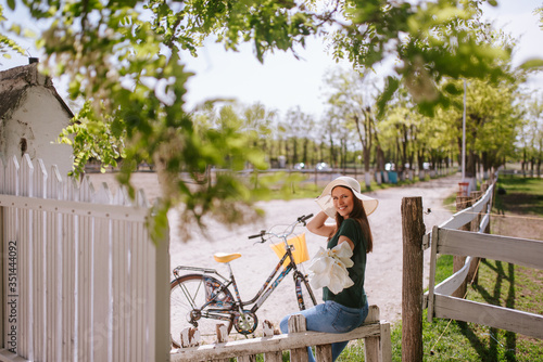 A young beautiful caucasian woman with a straw hat, bouquet of calla and a bicycle next to a fence in the village