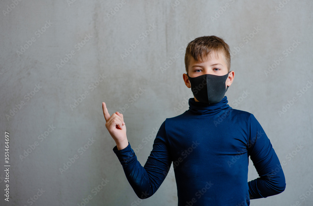 A boy in a black medical mask on his face on a uniform gray background points with his index finger to an empty place next to him and looks at the camera. Place for text A boy in a black medical mask on his face on a uniform gray background points with his index finger to an empty place next to him and looks at the camera. Place for text