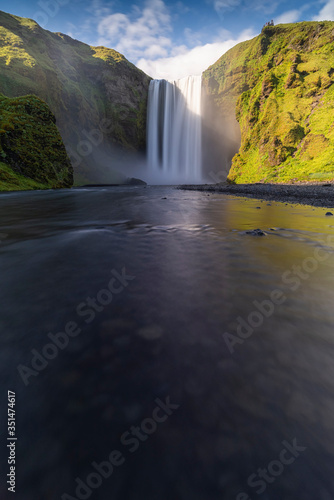 Beuatiful and powerful of streaming at Skogarfoss waterfall and orange sunlight cover green field around waterfall in evening,Summer,Iceland.