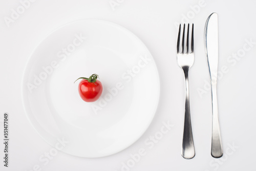 Eating on a low calorie concept. Top above overhead view photo of a plate with a single tomato with fork and knife placed to the right side isolated on white background