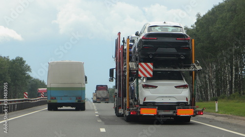 Loaded two-level car carrier truck with two-axle car transporter semi trailer drive on suburban highway road, rear view close up, new cars delivery logisticsat summer day on trees background