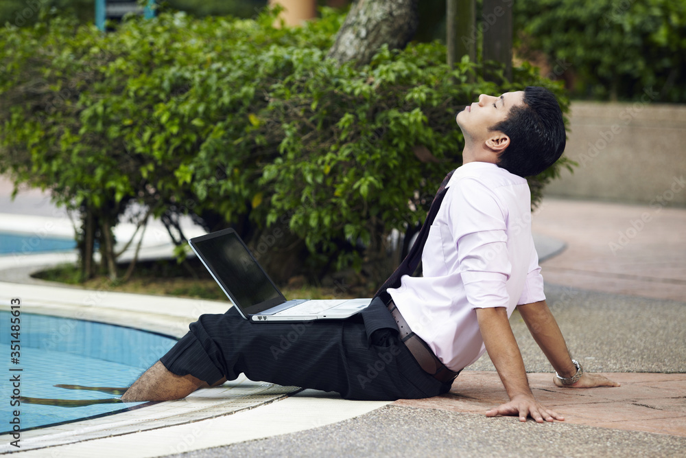 Young businessman relaxing by the pool with laptop on his lap