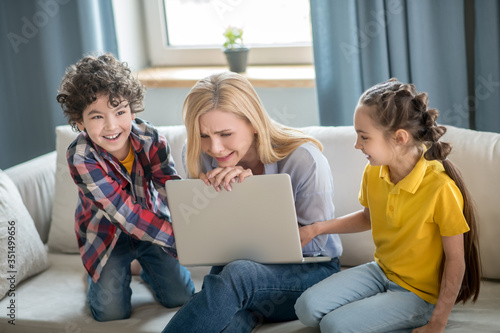 Distressed blonde female sitting on sofa, holding laptop on her knees, curly boy and dark-haired girl fighting over it © zinkevych