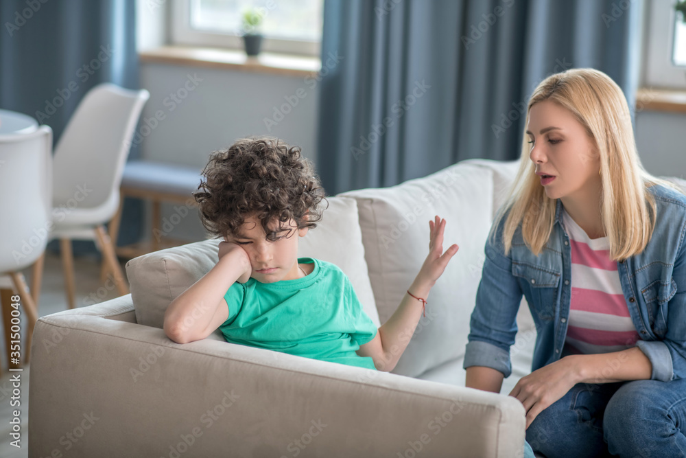 Sad curly boy sitting on sofa, blonde female talking to him, trying to cheer him up