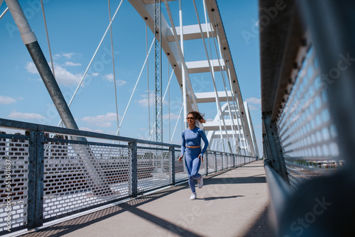 A young beautiful caucasian girl in blue sports equipment is running on the bridge. Training in nature