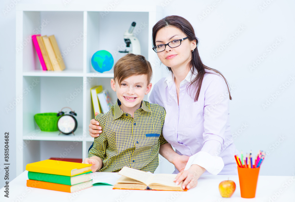 A boy sitting at a table at home in front of books and notebooks. A teacher sitting next to a boy helping him do homework A boy sitting at a table at home in front of books and notebooks. A teacher sitting next to a boy helping him do homework