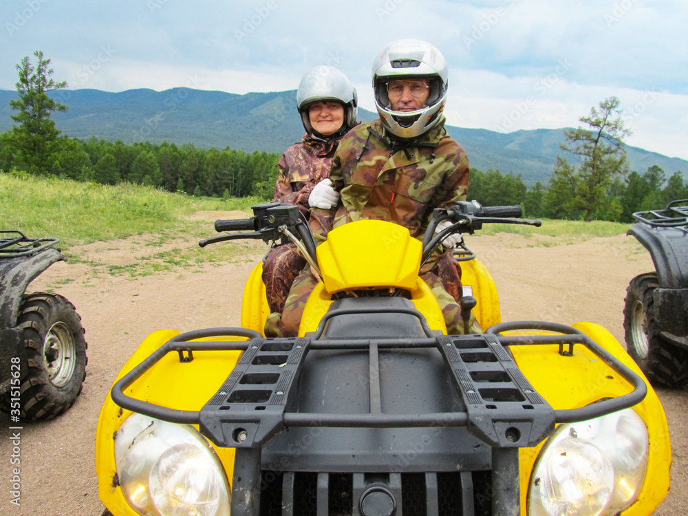 A man and woman of retirement age in camouflage suits and helmets ride a yellow ATV in the mountains. Sports, recreation, entertainment