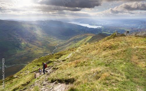 A hiker and their dog descending the summit of High Pike, Scandale beck below, Red Screes to the left and Lake Windermere in the distance on a sunny day in the Lake District.