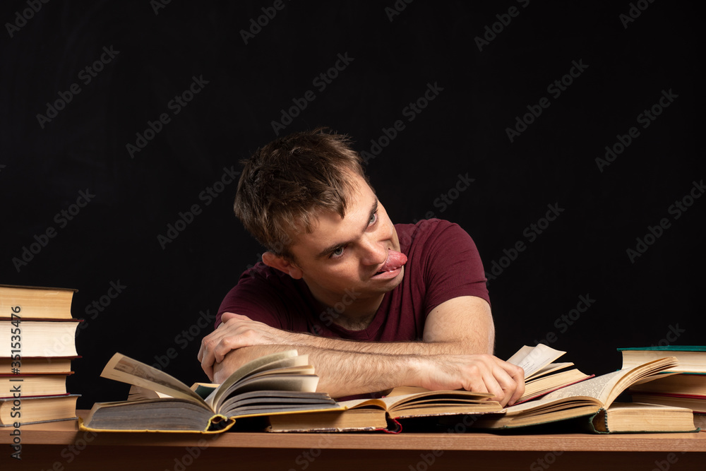 A young man was sitting at a table above books, holding his head in his hands. Piles of books are lying nearby. Black background. isolate. difficulties in education and quarantine. A young man was sitting at a table above books, holding his head in his hands. Piles of books are lying nearby. Black background. isolate. difficulties in education and quarantine.