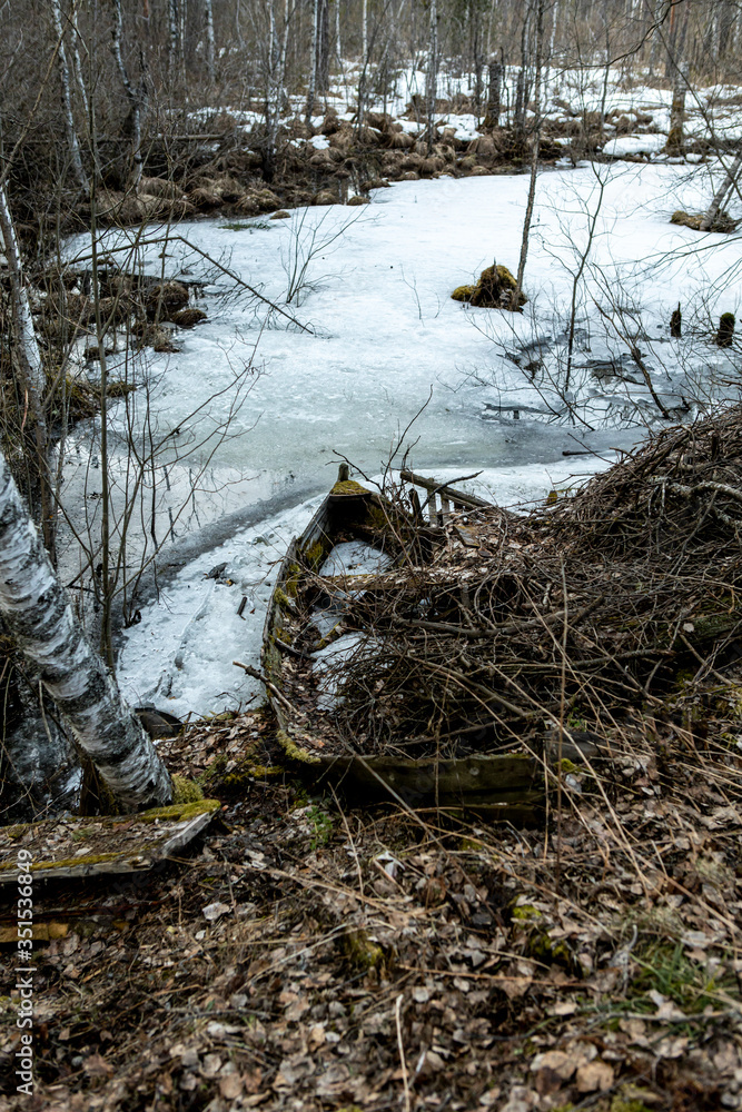 Obraz premium old wooden boat tied at the edge of a forest lake covered with snow and ice.