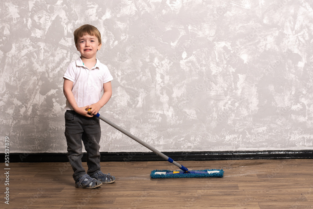 a little boy in jeans and a white T-shirt is standing in the apartment with a mop in his hands. the child is crying and does not want to help parents clean the apartment. spring-cleaning. vagaries in a little boy in jeans and a white T-shirt is standing in the apartment with a mop in his hands. the child is crying and does not want to help parents clean the apartment. spring-cleaning. vagaries in