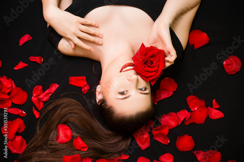 Portrait of a young, attractive woman with a rose in her hands lying on rose petals on a black background. Valentine's day concept. Model with retouched skin.