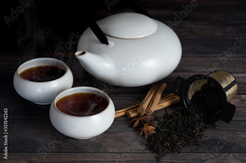 two cup of tea, teapot, cinnamon stick, tea strands, strainer, star anise on wooden table.