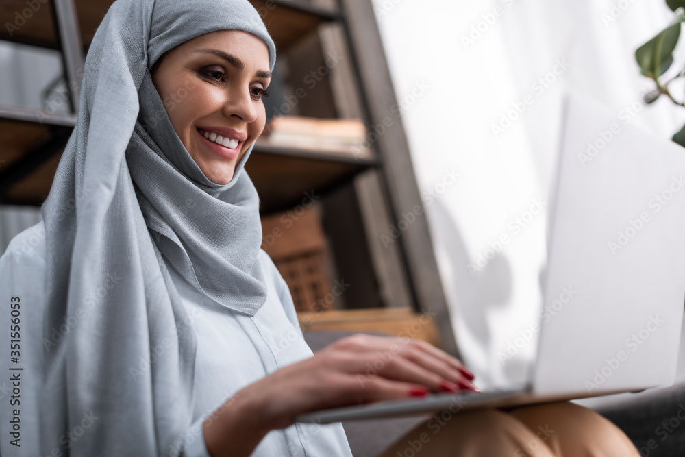 Selective focus of smiling Arabian woman in hijab using laptop in living room Selective focus of smiling Arabian woman in hijab using laptop in living room